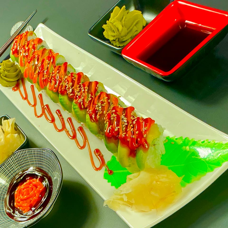 Sushi roll with avocado, red sauce, sesame seeds, wasabi, and soy sauce on a white plate.