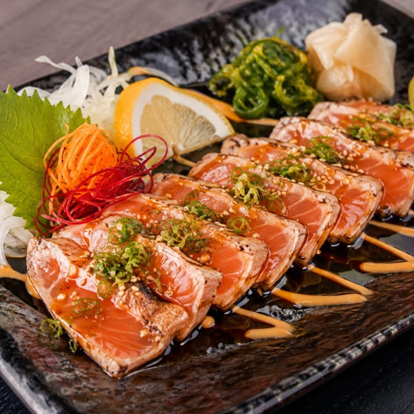 Seared salmon sashimi with green onions, sesame seeds, lemon, seaweed, and ginger on a black plate.